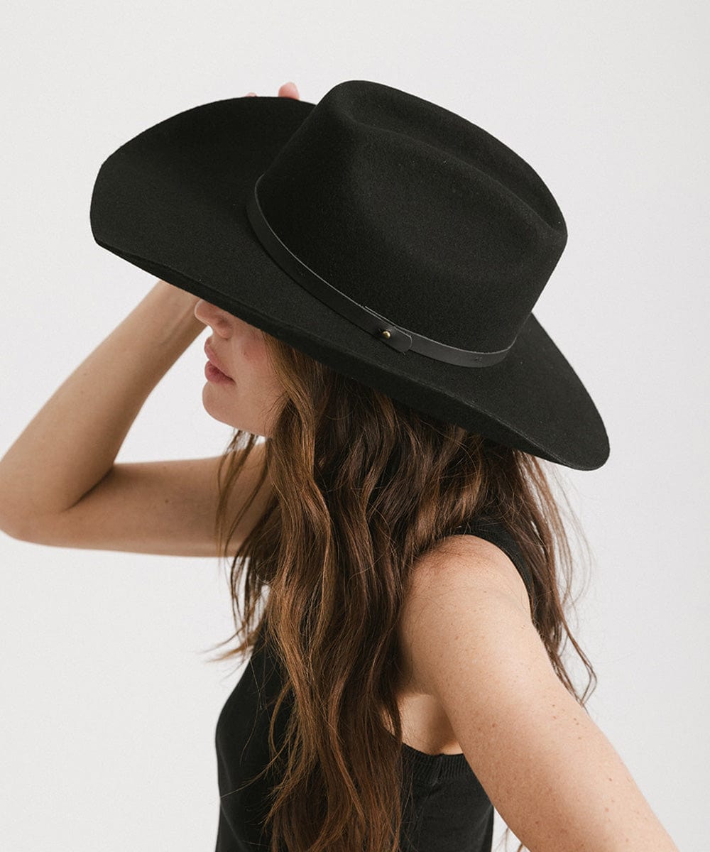 Woman wearing a black cowboy hat with an upturned brim and an attached leather band and a black top against a white background #color_black