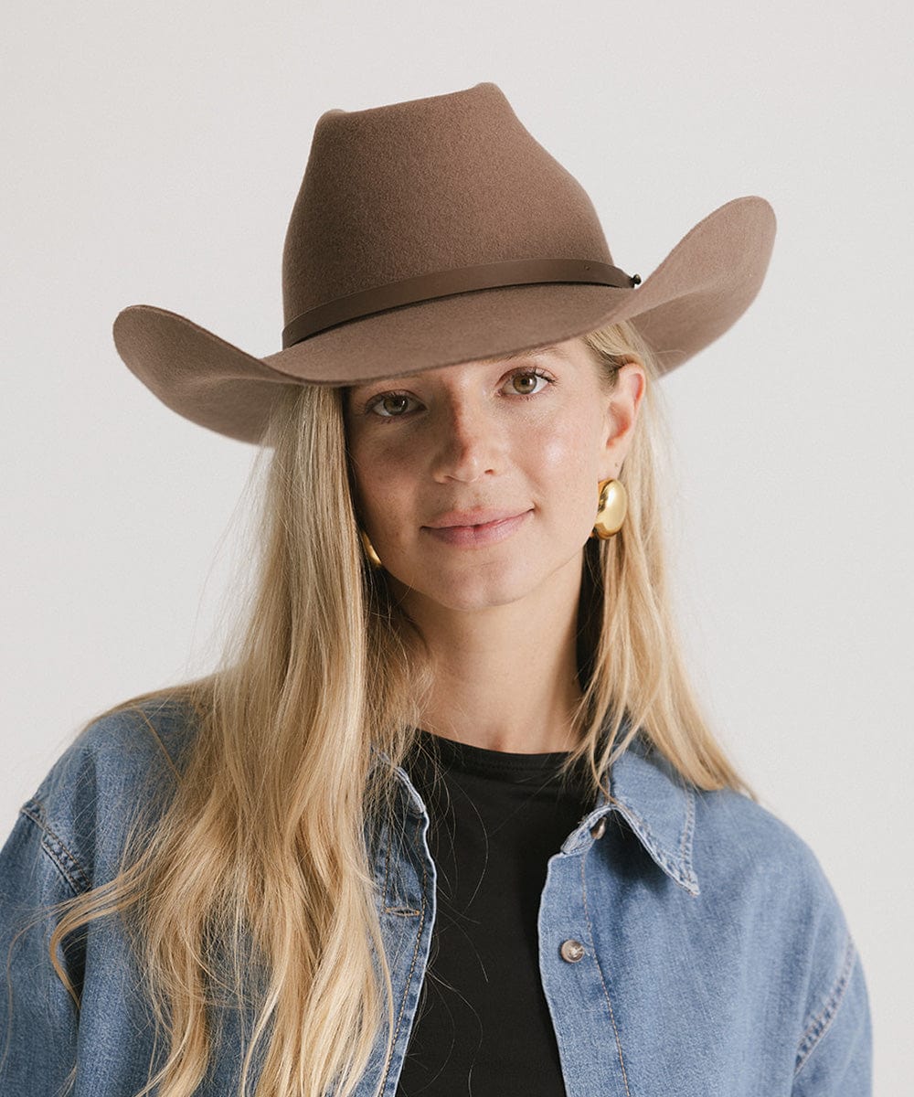Woman wearing a brown cowboy hat with an upturned brim and an attached leather band, and a denim jacket against a white background #color_chocolate