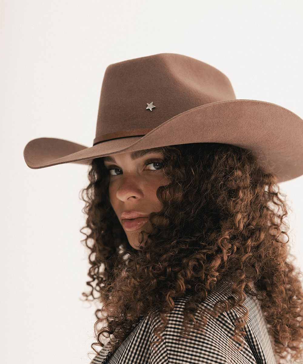 Woman wearing a chocolate colored cowboy hat with an upturned brim and an attached leather band with a star pin, displayed against a white background. #color_chocolate