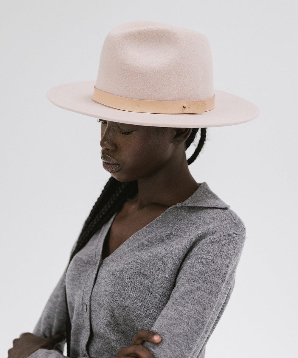 Woman wearing a blush pink colored fedora hat with a hat band and a grey shirt, posing sideways against a plain background  #color_blush pink