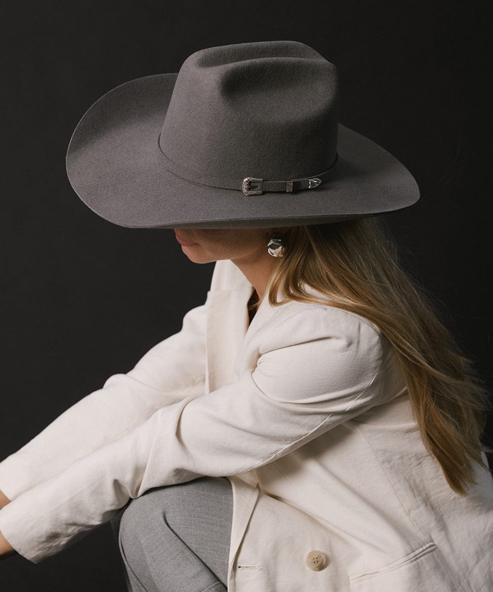 Woman wearing a limited edition stone colored cowboy hat with a hat band, a white blazer and grey pants, posing against a dark grey background #color_limited edition stone