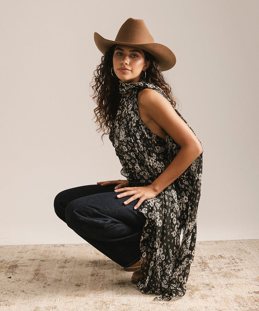 Woman wearing a caramel colored cowboy hat, black floral halter top, and dark blue jeans, sitting against a plain background #color_caramel