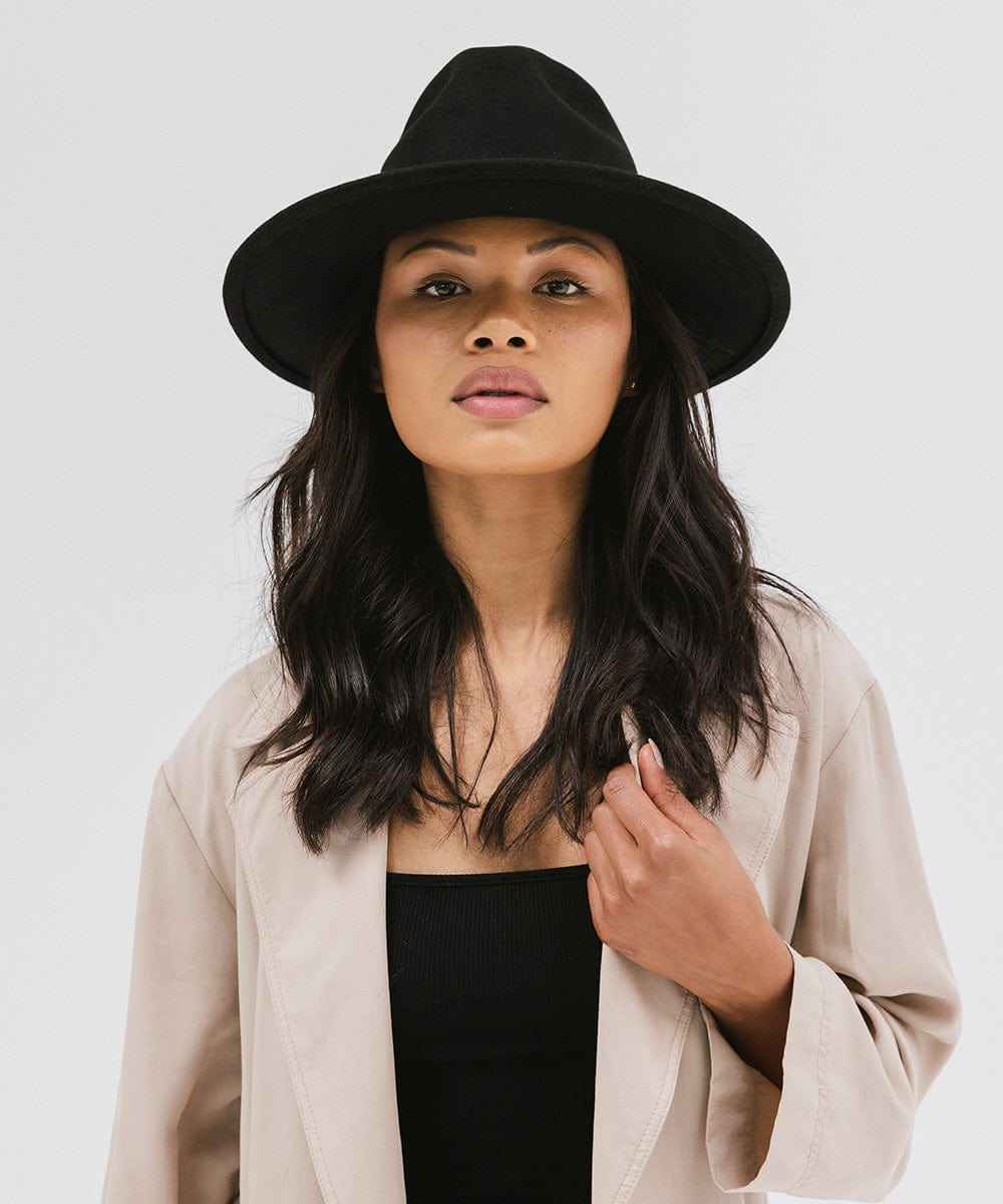 Woman wearing a black fedora hat and a beige coat with a black top  against a plain background #color_black