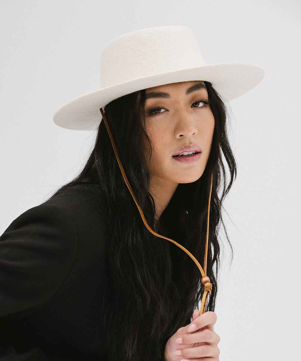 Woman wearing an off white wide-brimmed hat with a strap and a black coat, posing sideways against a plain background #color_off white