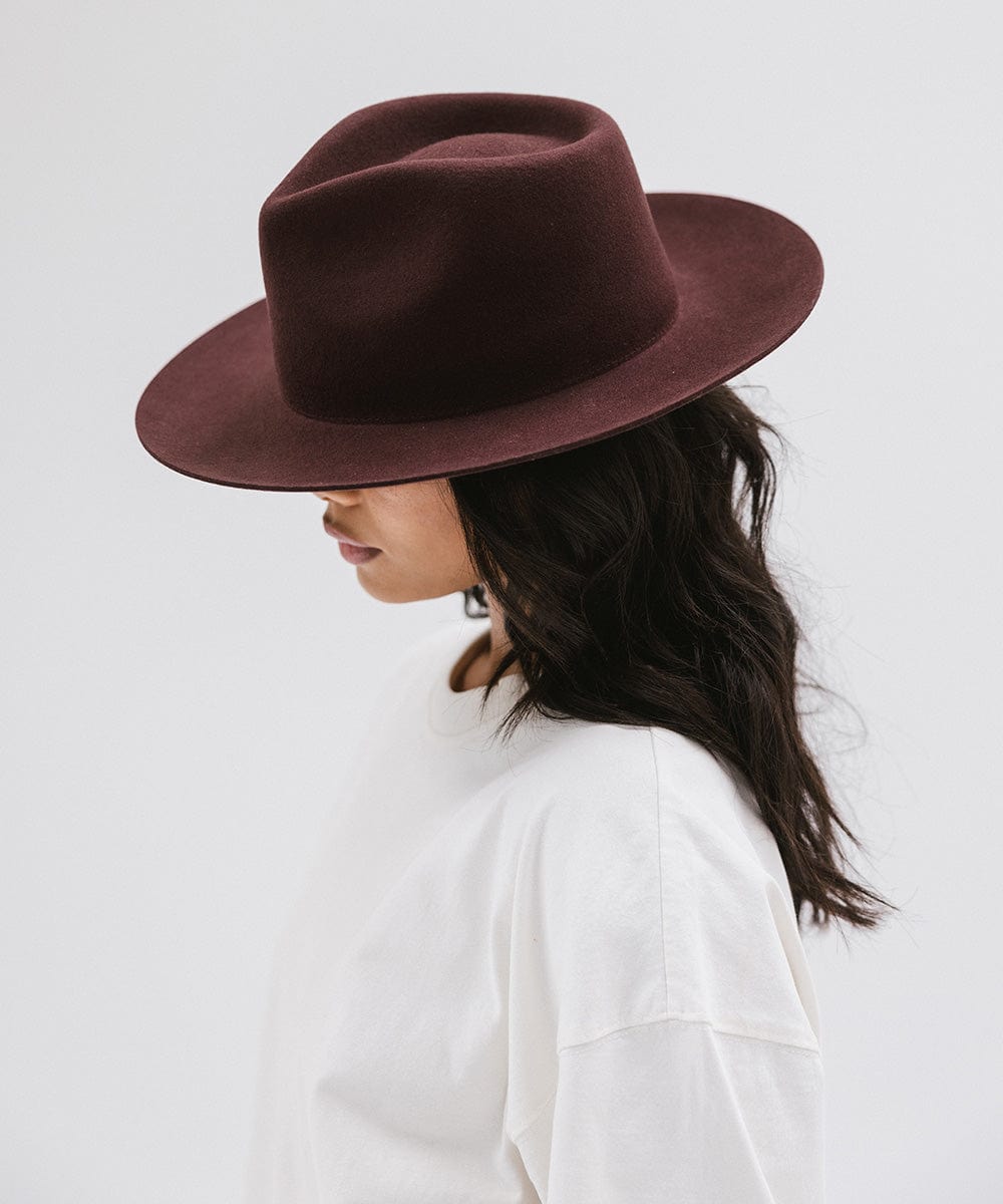 Woman wearing a dark cherry colored fedora hat and a white shirt, facing sideways against a white background #color_dark cherry