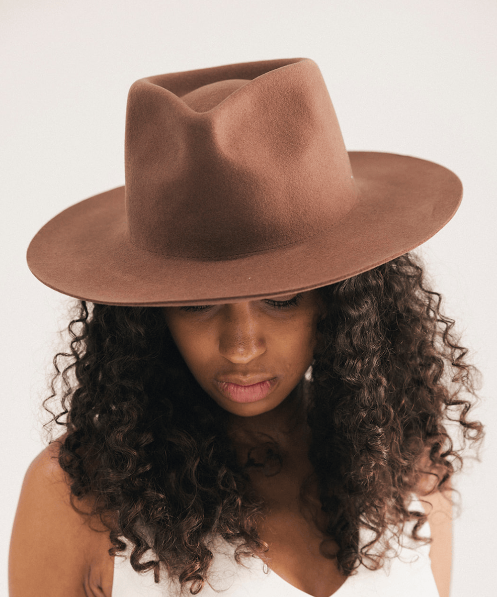 A woman with long, curly dark brown hair wearing a brown felt fedora hat, looking down against a plain light background #color_dark oak
