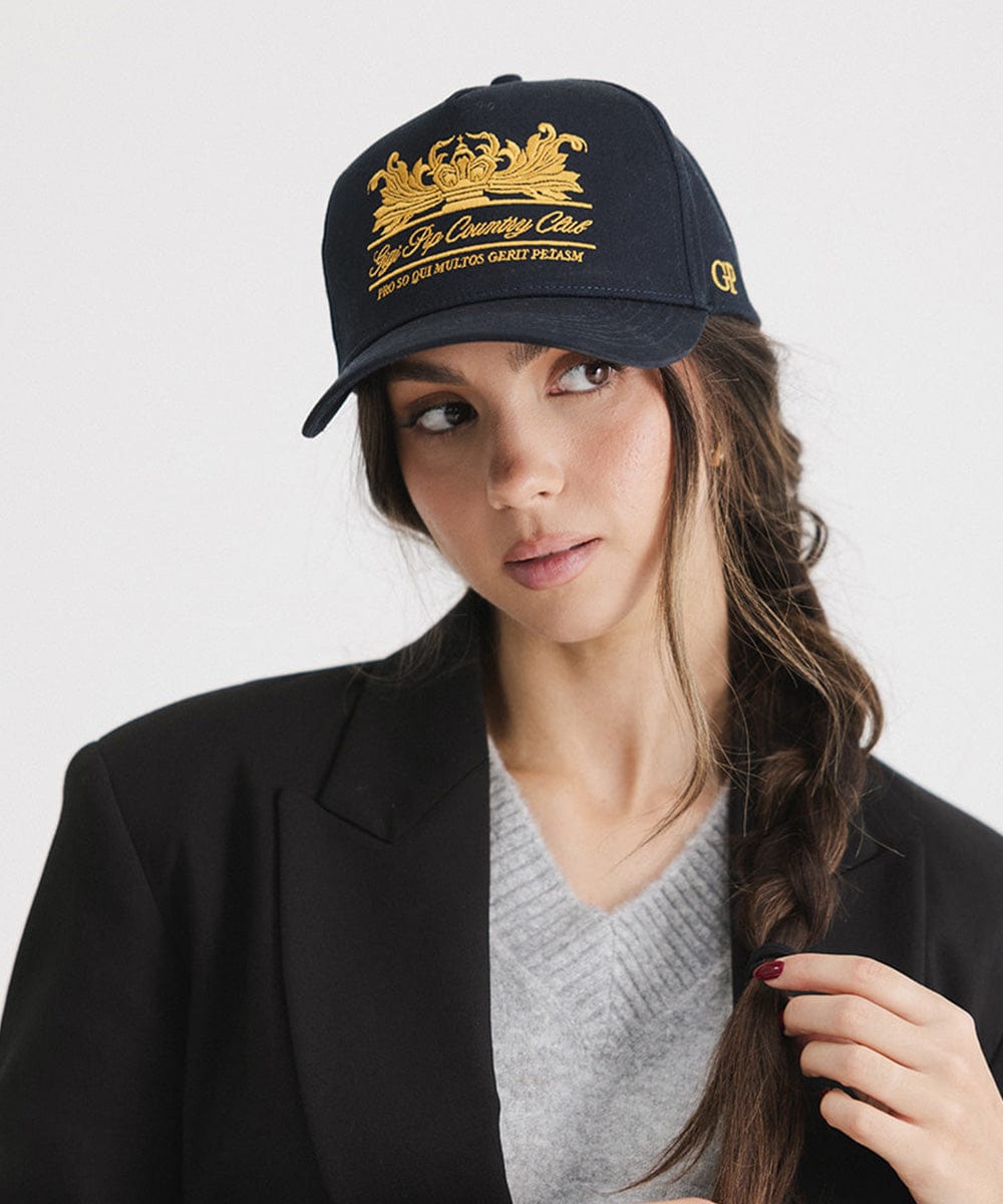 Woman wearing a navy vintage trucker cap with a gold logo and a grey top with a black blazer against a plain background #color_navy