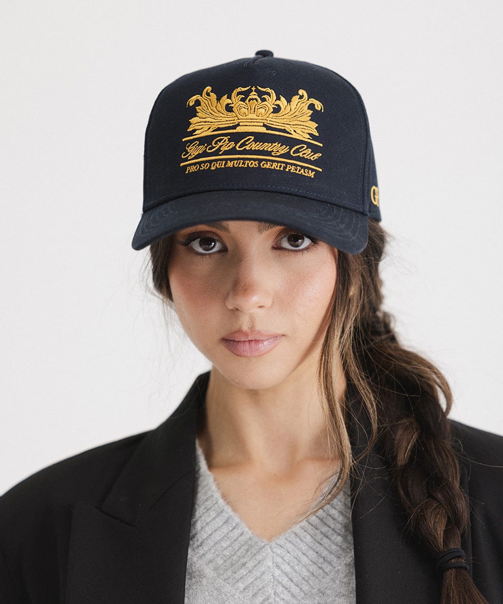 Woman wearing a navy vintage trucker cap with a gold logo and a black blazer against a plain background #color_navy