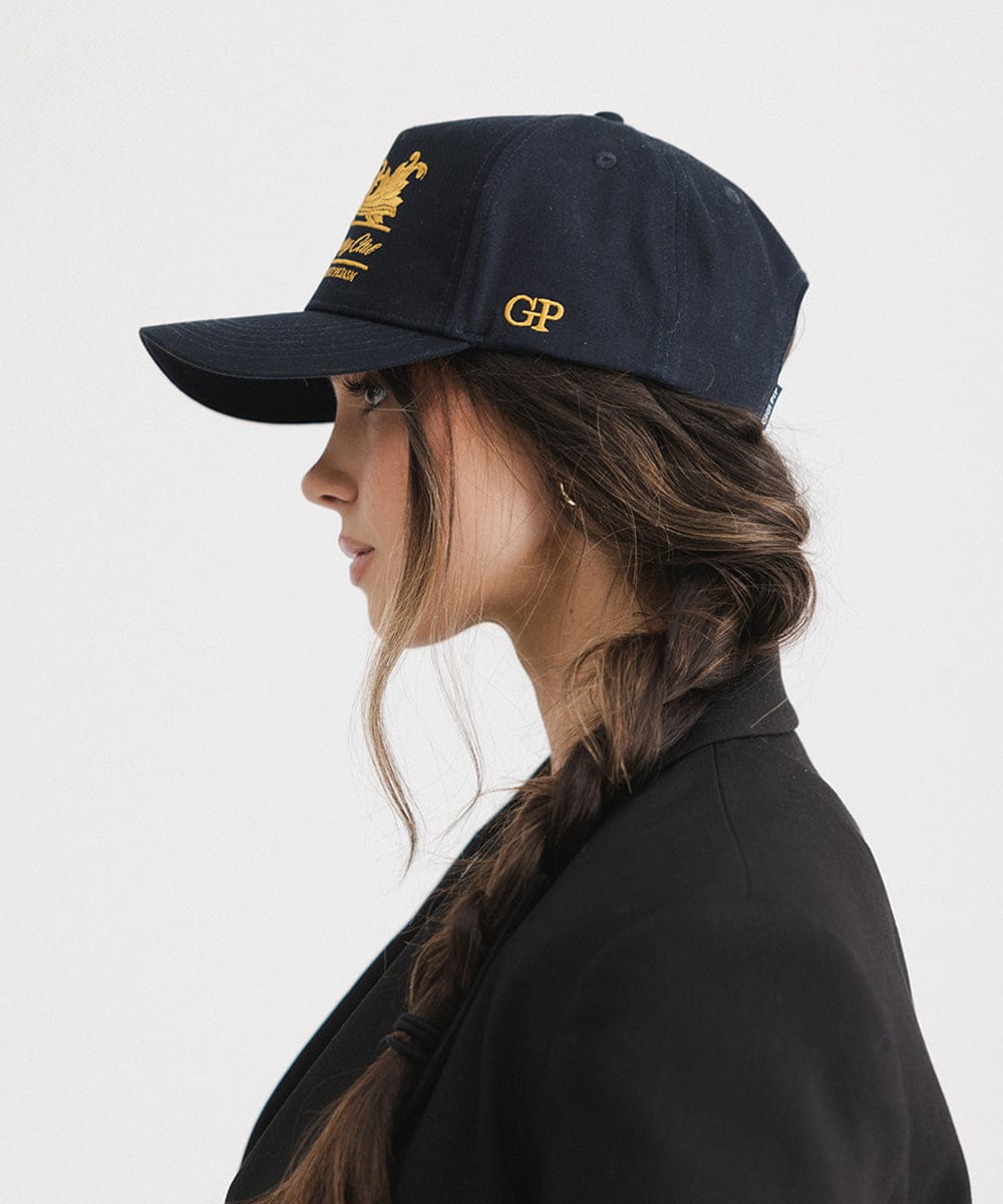Woman wearing a navy vintage trucker cap with a gold logo, and a black blazer, facing sideways against a plain background #color_navy
