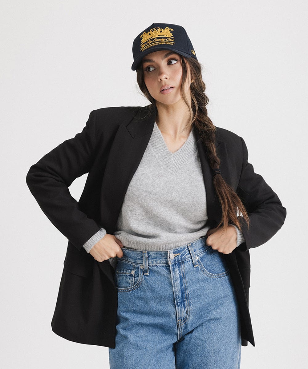 Woman wearing a navy vintage trucker cap with a gold logo, a grey top with a black blazer and blue jeans against a plain background #color_navy