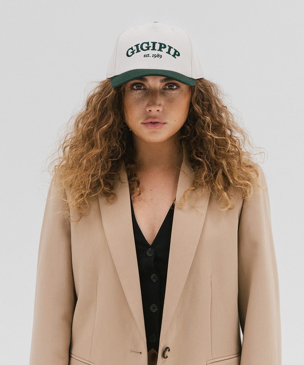 Woman wearing a cream and dark green trucker hat with 'GIGI PIP' embroidered in dark green on the front and a beige coat against a plain background #color_cream - dark green