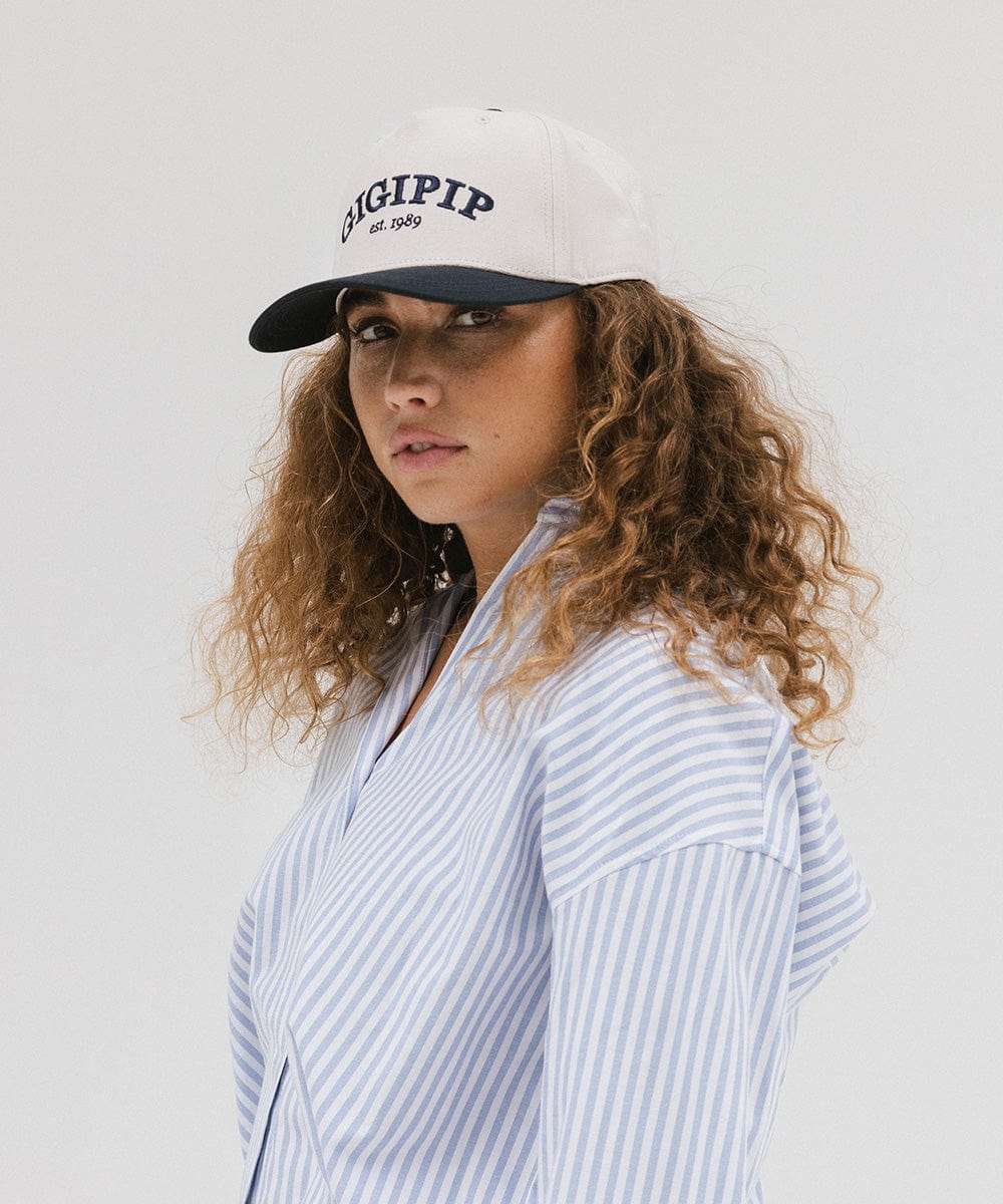 Woman wearing a cream and navy trucker hat with 'GIGI PIP' embroidered in navy on the front and a light colored shirt, posing sideways against a plain background #color_cream - navy