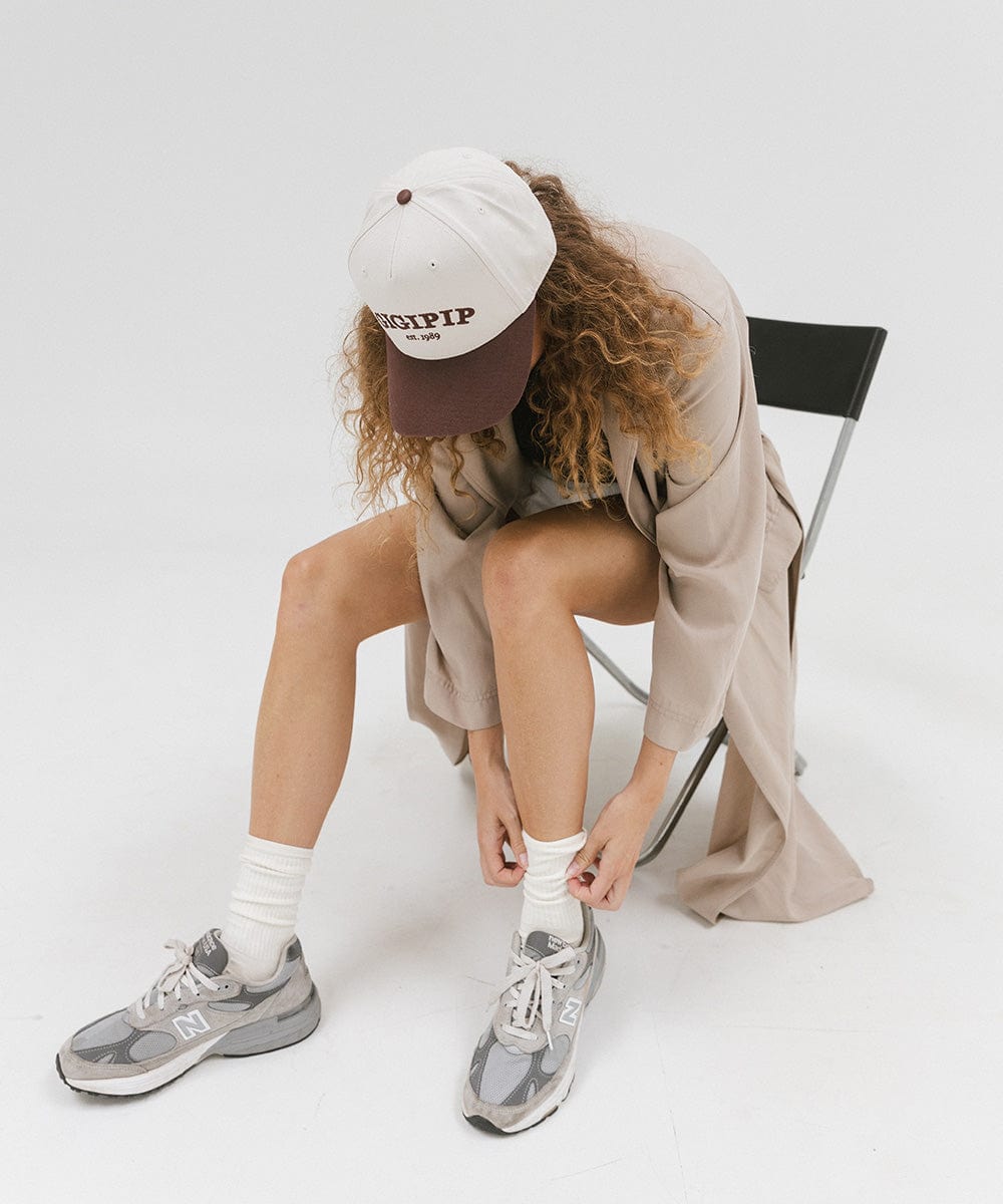 Woman wearing a cream and chocolate brown trucker hat with 'GIGI PIP' embroidered in chocolate brown on the front, a beige coat and grey shoes, sitting on a chair against a plain background #color_cream - chocolate brown