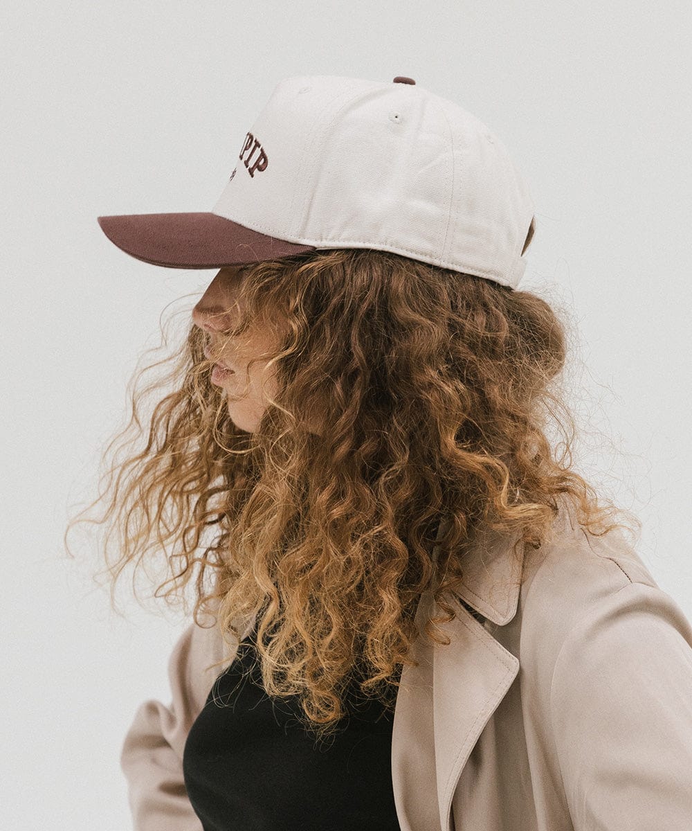 Woman wearing a cream and chocolate brown trucker hat and a black top with a beige coat, facing sideways against a plain background #color_cream - chocolate brown