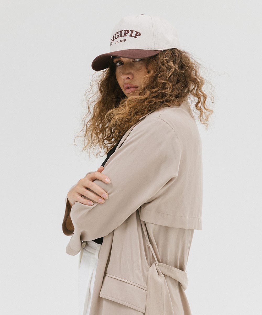 Woman wearing a cream and chocolate brown trucker hat with 'GIGI PIP' embroidered in chocolate brown on the front and a beige coat, posing sideways against a plain background #color_cream - chocolate brown