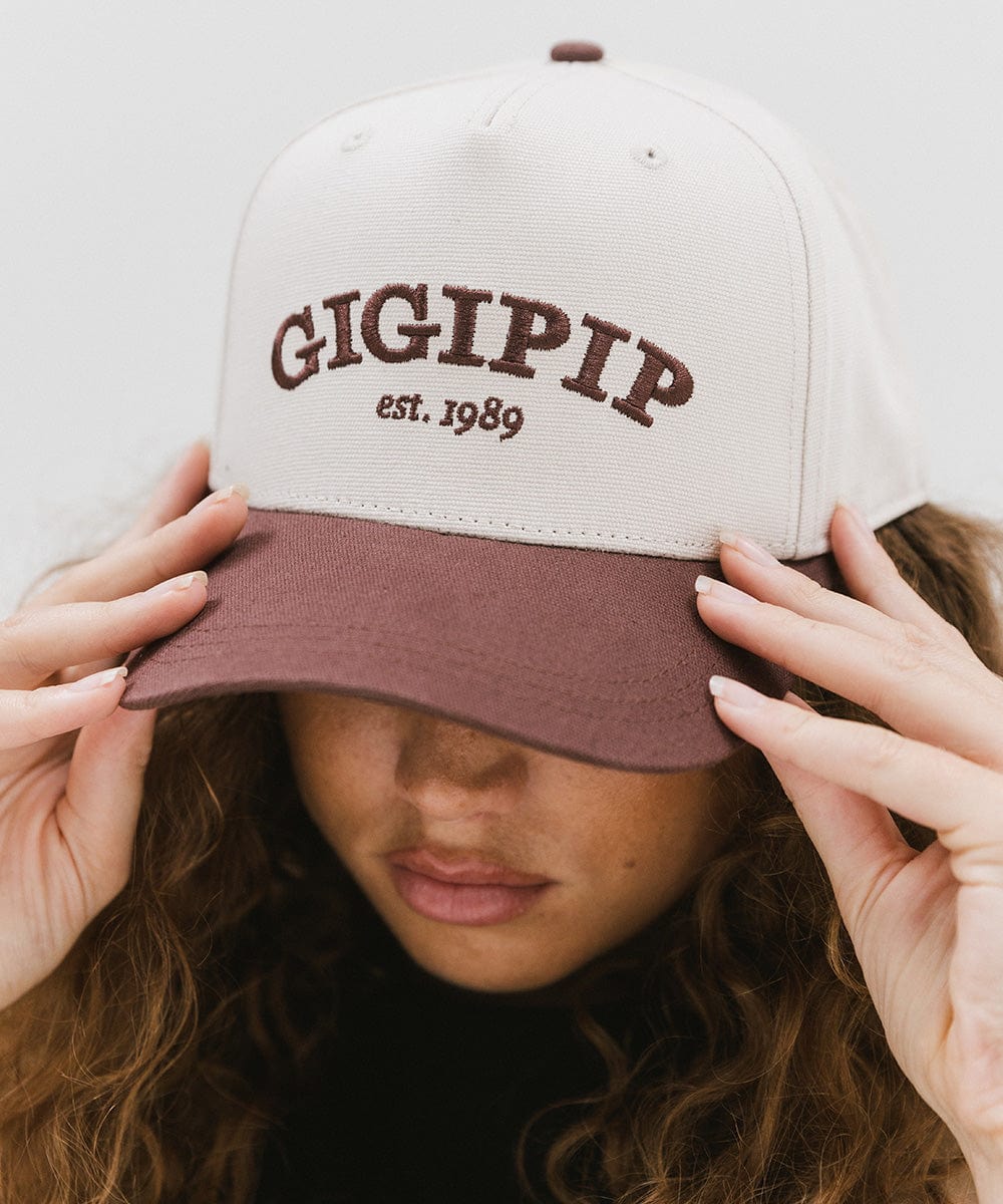Woman wearing a cream and chocolate brown trucker hat with 'GIGI PIP' embroidered in chocolate brown on the front against a plain background #color_cream - chocolate brown