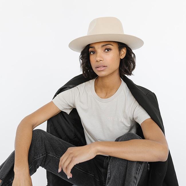 Woman wearing a sand colored t-shirt with 'GIGI PIP' text, a cream hat, black coat and dark grey jeans against a white background #color_sand