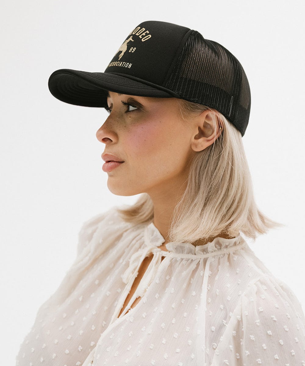 Woman wearing a black cap with golden 'GIGIRODEO' text and logo, and a white top, facing sideways against a plain background #color_black