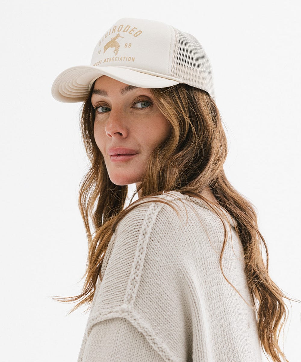 Woman wearing a cream cap with golden 'GIGIRODEO' text and logo, and a grey sweater against a plain background #color_cream