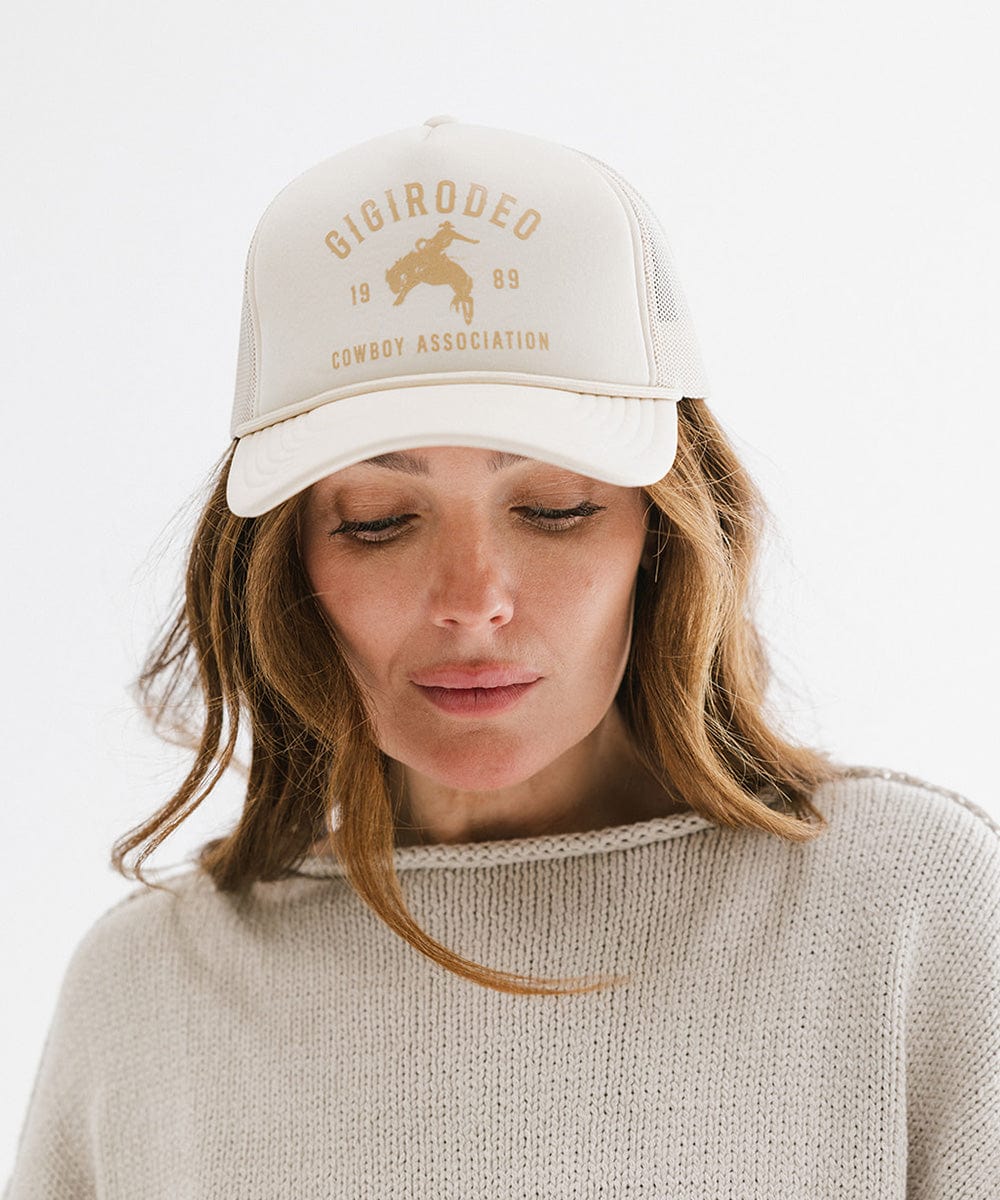 Woman wearing a cream cap with golden 'GIGIRODEO' text and logo, and a grey sweater against a plain background #color_cream