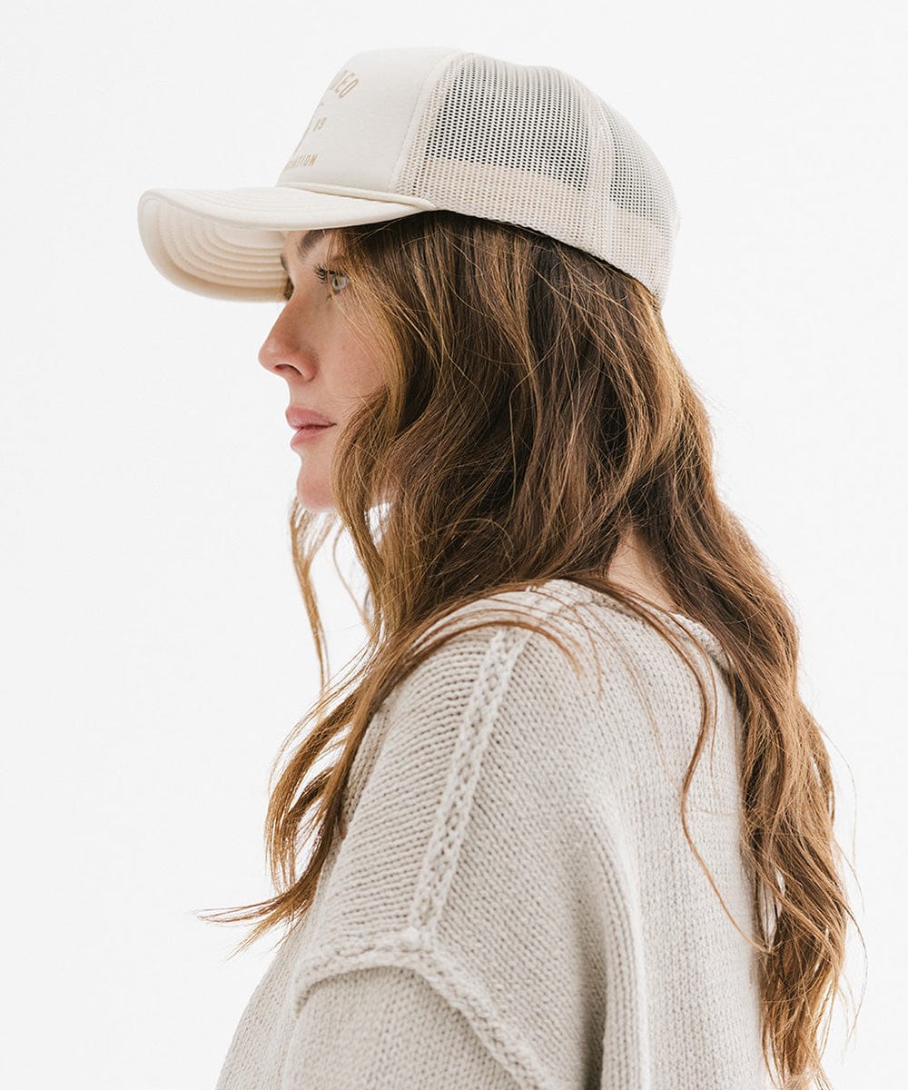 Woman wearing a cream cap with golden 'GIGIRODEO' text and logo, and a grey sweater, facing sideways against a plain background #color_cream