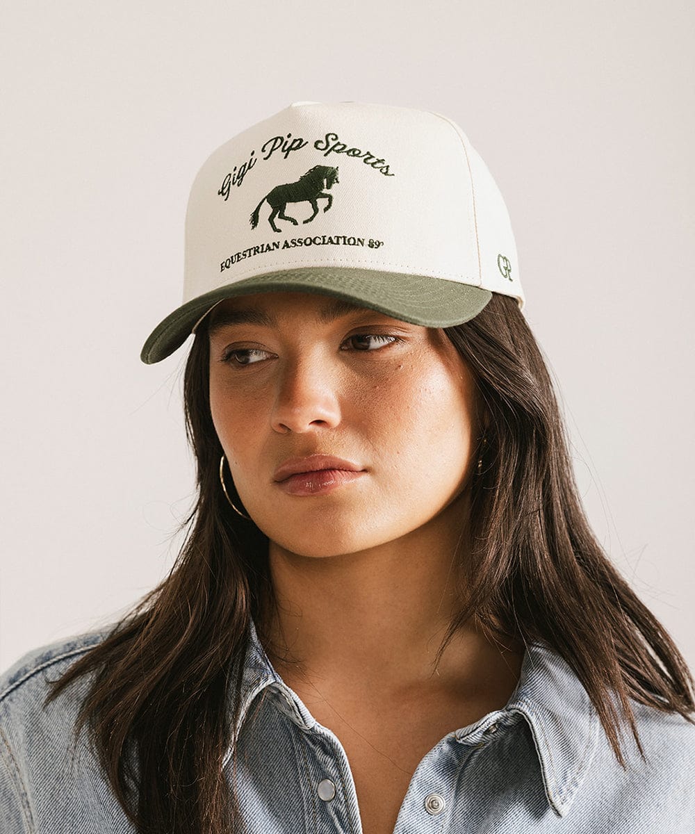 Woman wearing a cream and dark green Trucker hat with 'Gigi Pip Sports' and horse logo and a denim shirt, posing against a plain background #color_cream - dark green