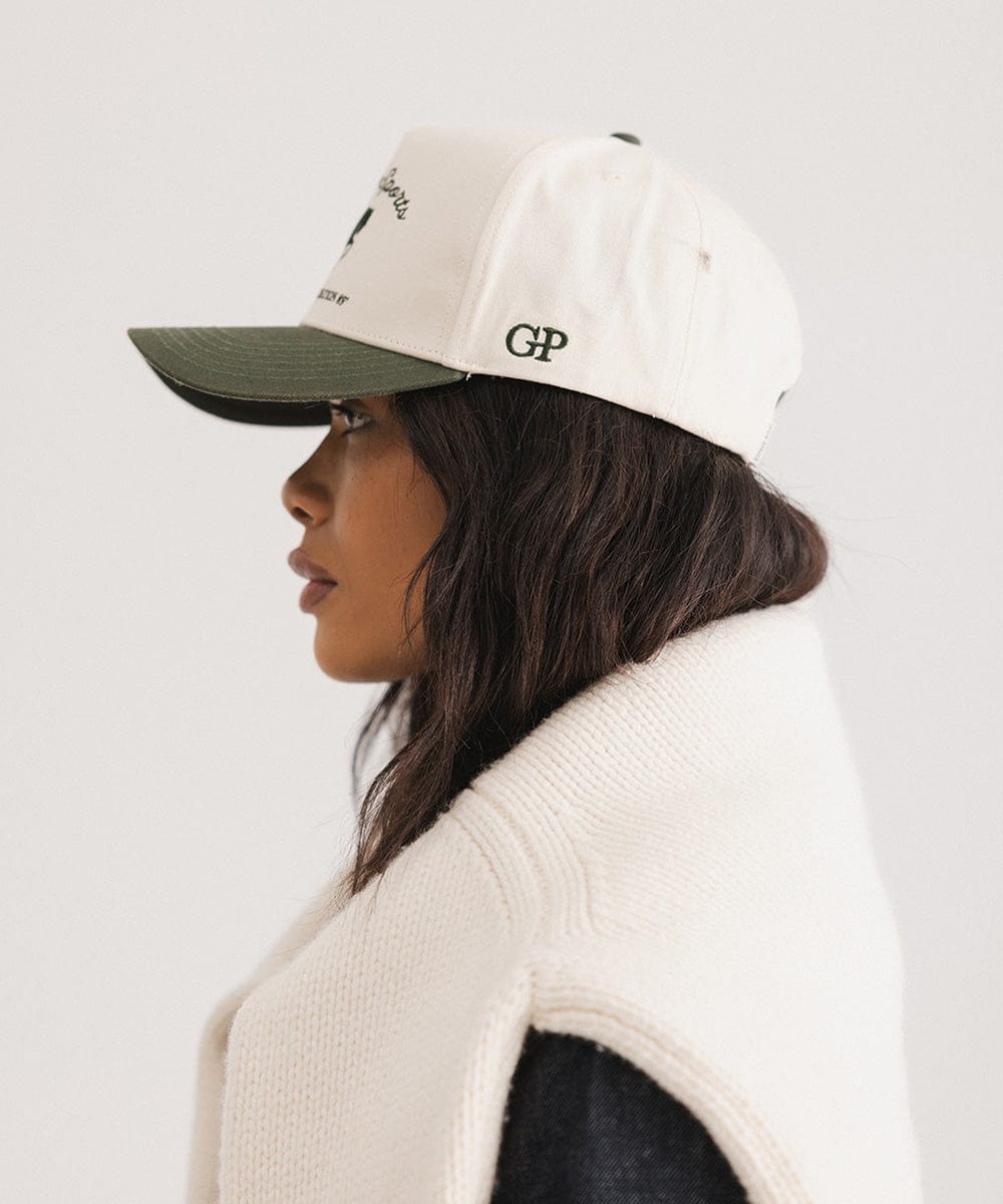 Woman wearing a cream and dark green Trucker hat and a white sweater, facing sideways against a plain background #color_cream - dark green
