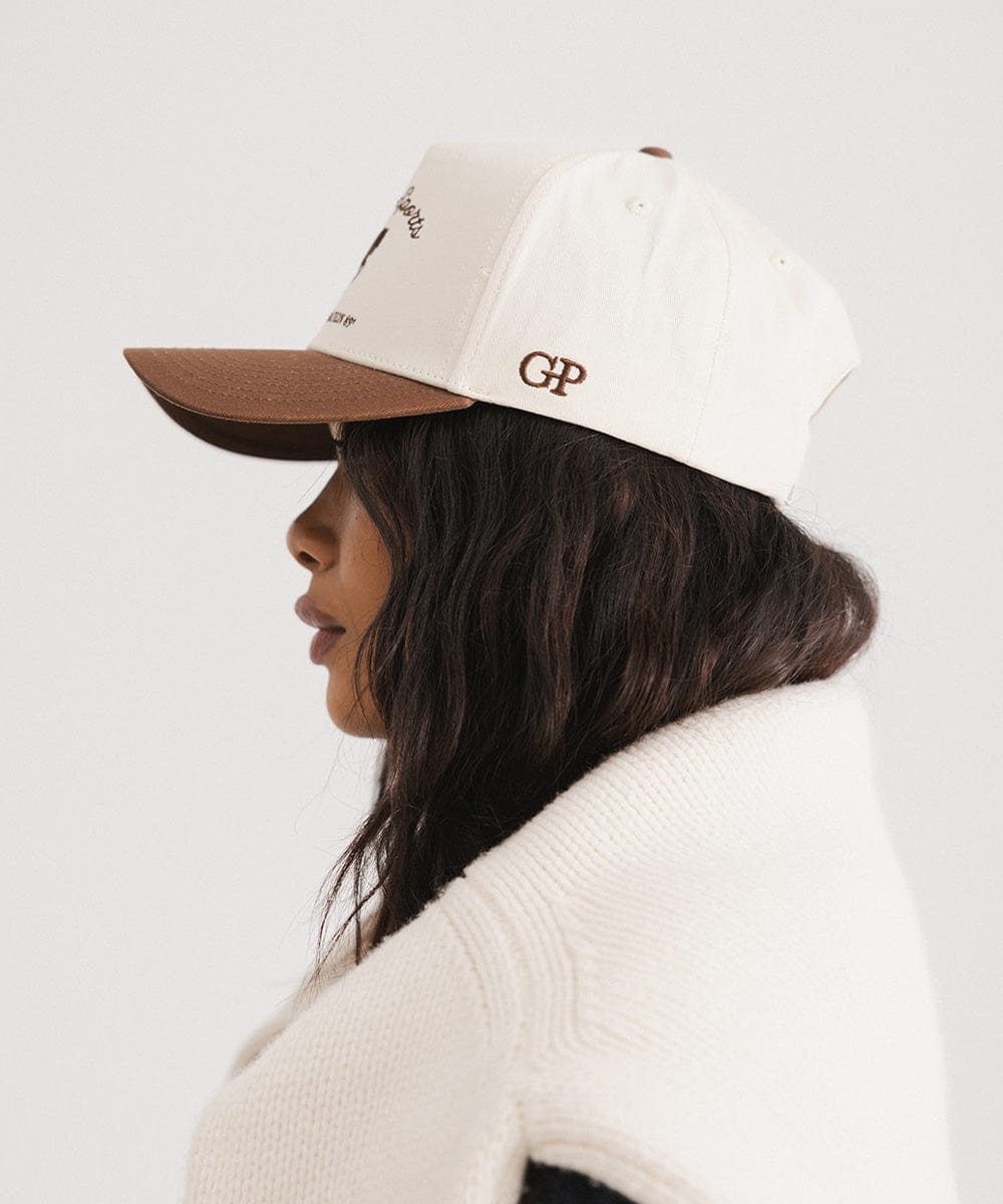 Woman wearing a cream and chocolate brown Trucker hat and a white sweater, facing sideways against a plain background #color_cream - chocolate brown