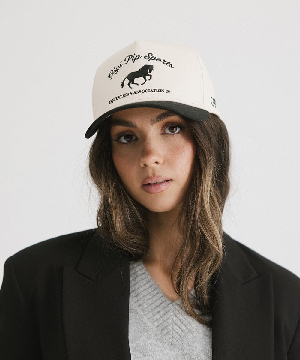 Woman wearing a cream and black Trucker hat with 'Gigi Pip Sports' and horse logo and a black blazer against a plain background #color_cream - black