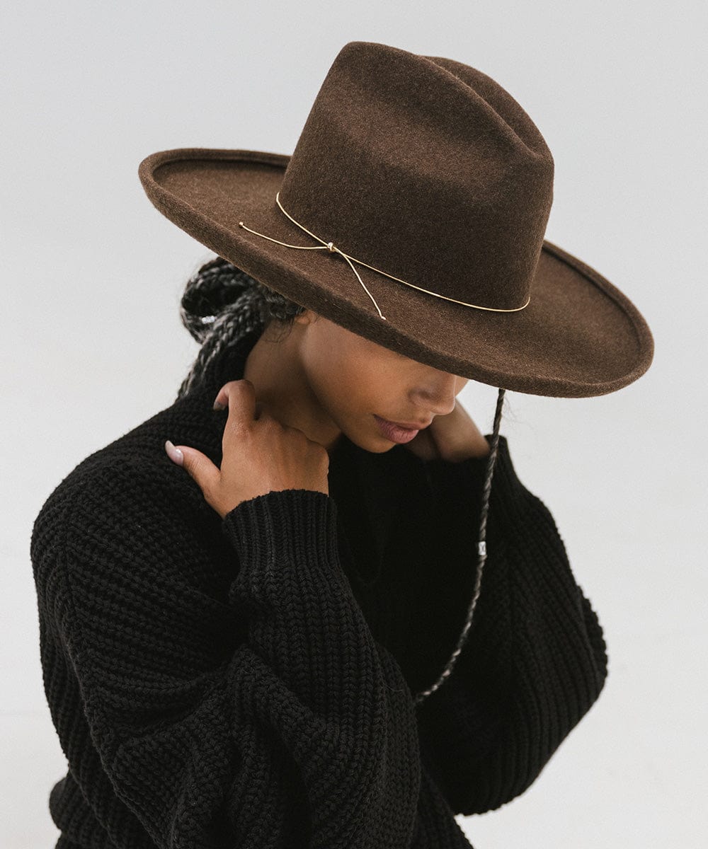 Woman wearing a brown hat with a gold chain bolo hat band and a black sweater, posing sideways against a plain background #color_gold