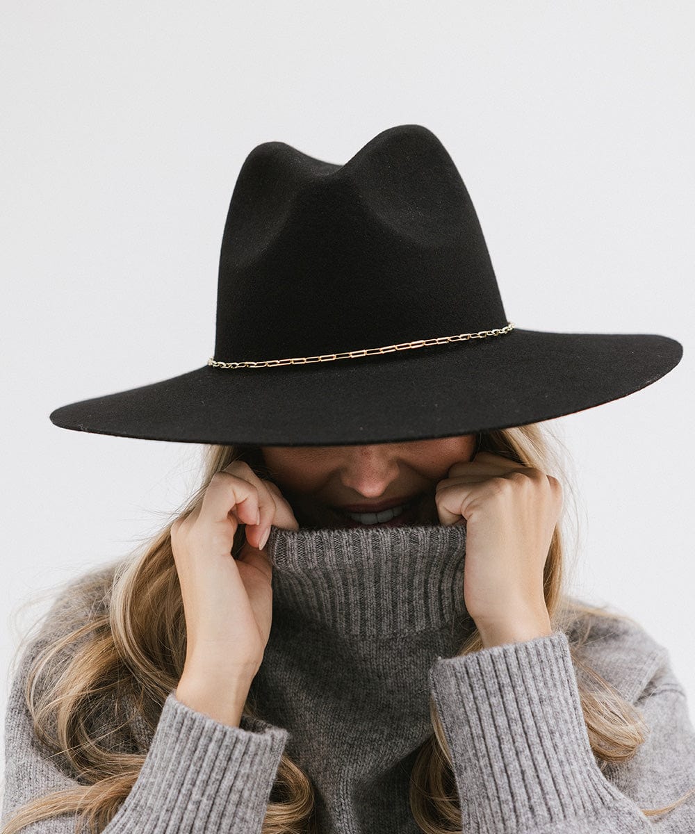 Woman wearing a black wide-brimmed hat with a gold paperclip chain hat band and a grey sweater, posing against a plain background #color_gold