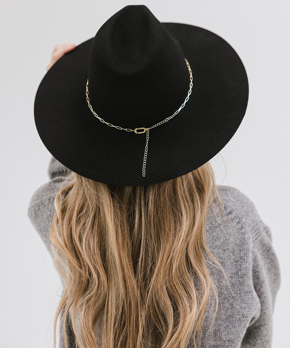 Woman wearing a black wide-brimmed hat with a gold paperclip chain hat band and a grey sweater, facing behind against a plain background #color_gold