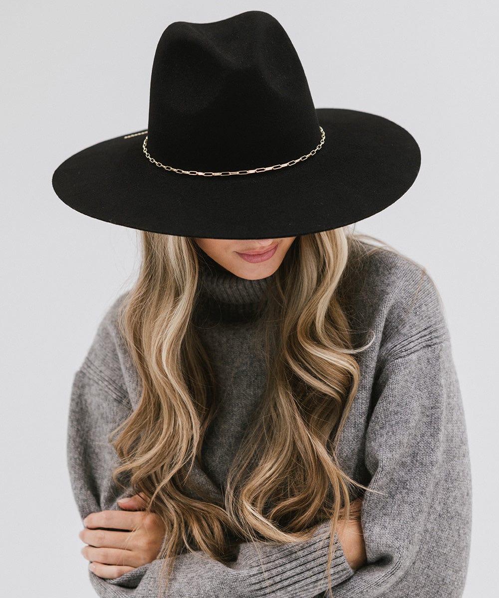 Woman wearing a black wide-brimmed hat with a gold paperclip chain hat band and a grey sweater against a plain background #color_gold