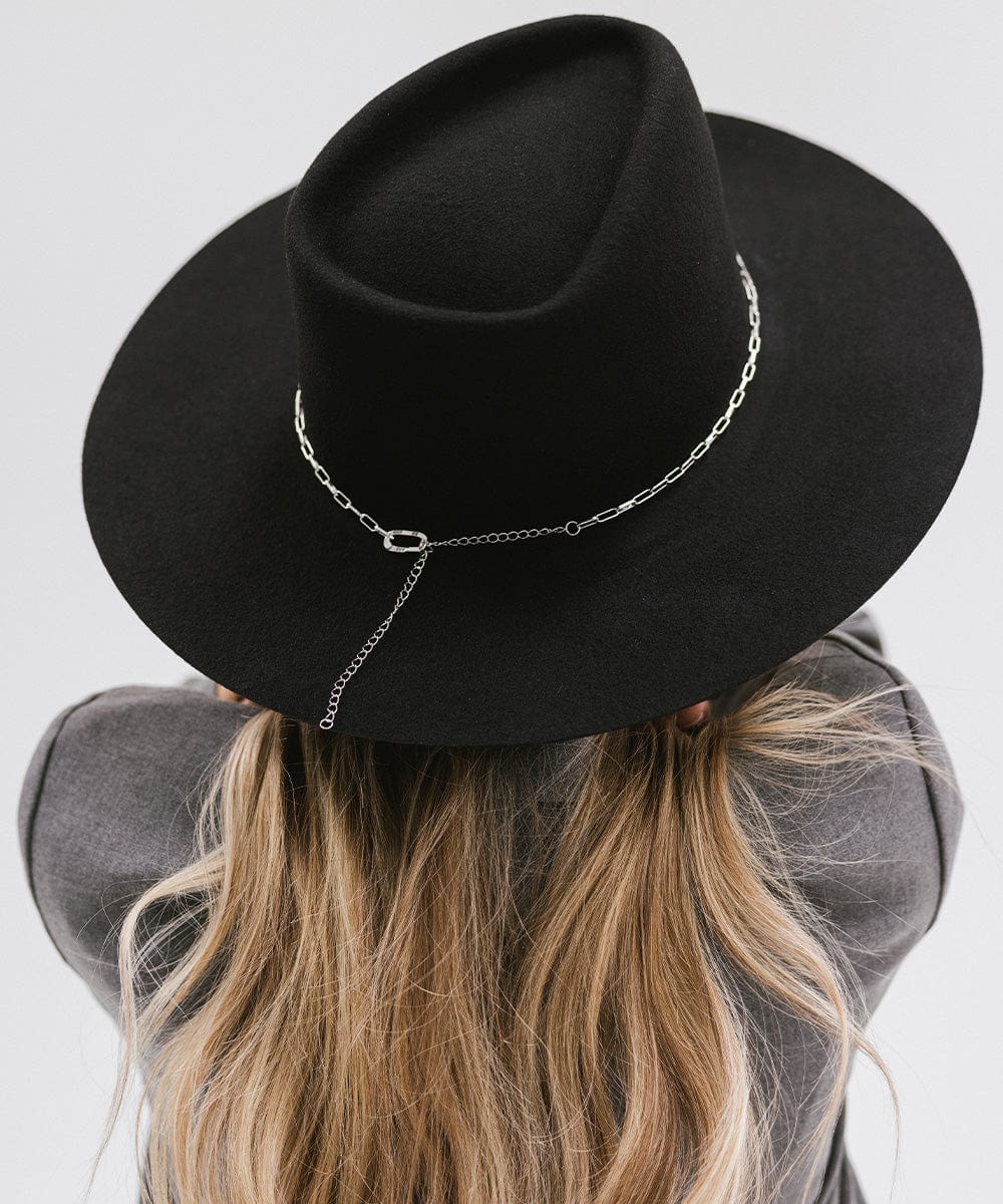 Woman wearing a black wide-brimmed hat with a silver paperclip chain hat band and a grey coat, facing behind against a plain background #color_silver