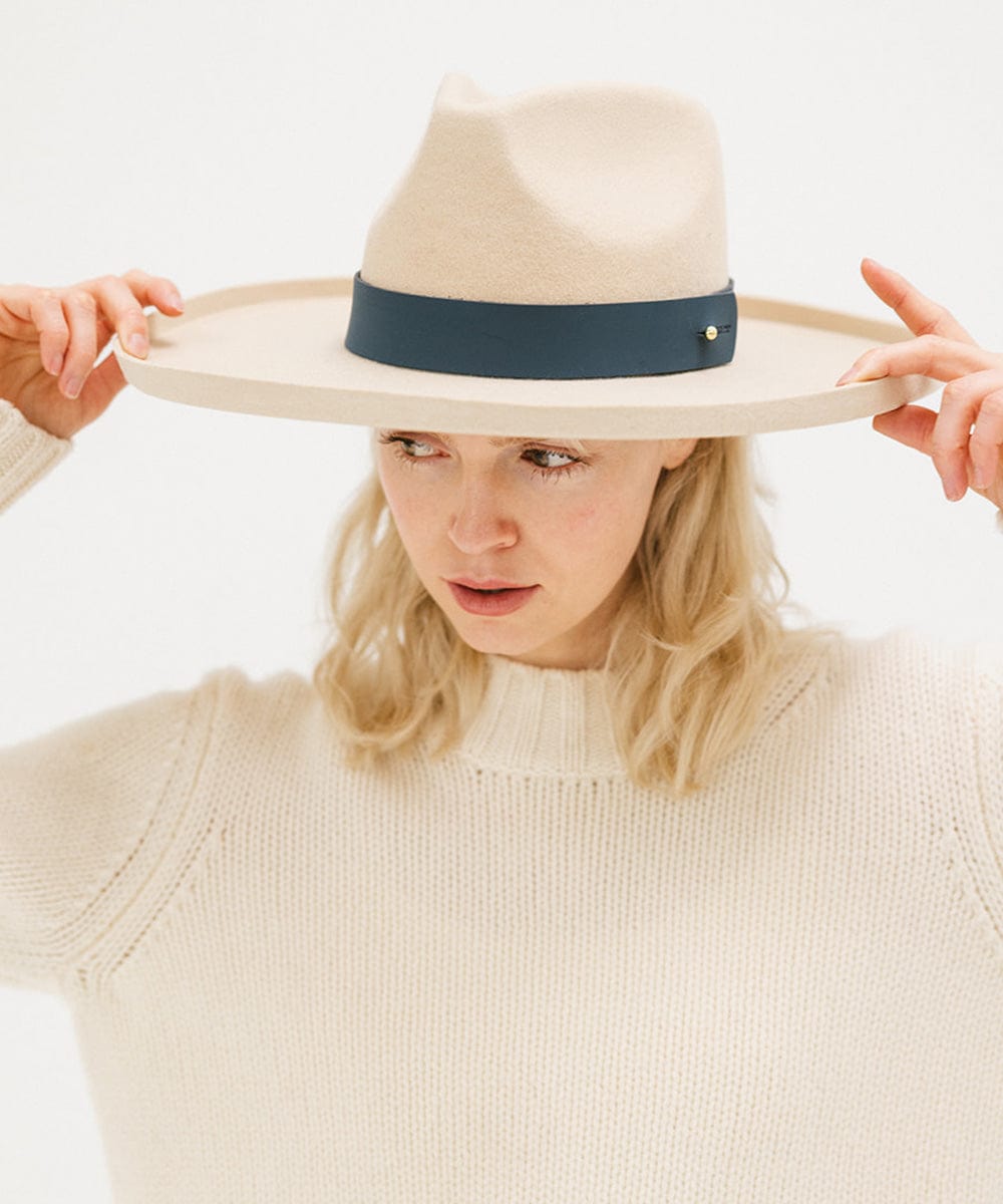 A woman wearing a cream hat with a wide vintage blue colored leather band around the base, and a cream sweater, posing against a plain background #color_vintage blue