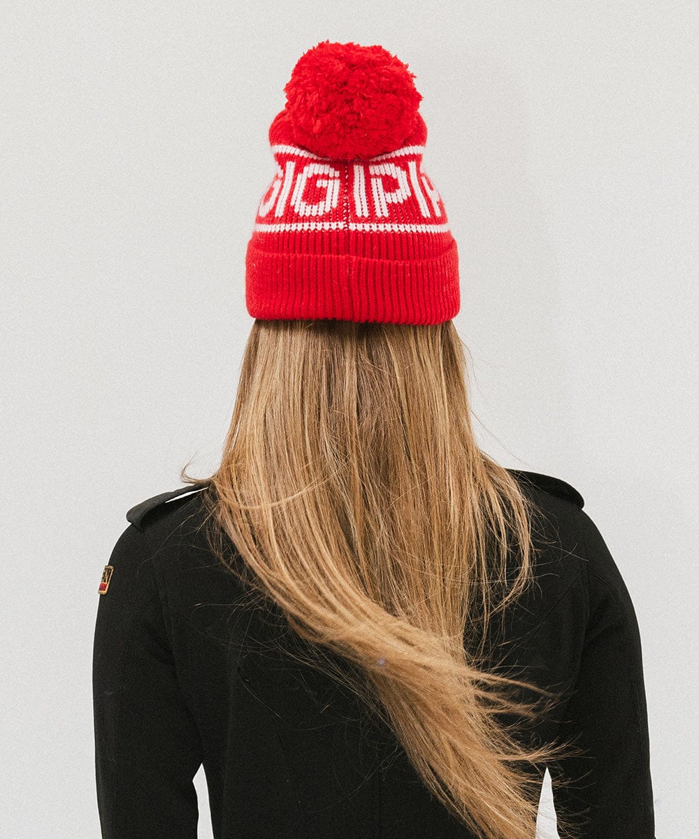 Woman wearing a ruby red colored knit beanie with pom-pom and 'GIGI PIP' branding and a black jacket, facing behind against a plain background #color_ruby red