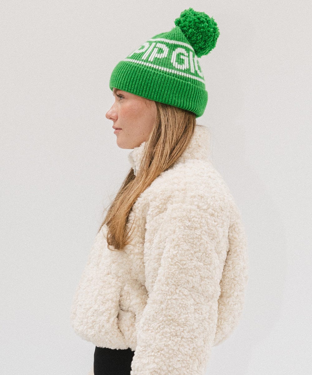 Woman wearing an evergreen colored knit beanie with pom-pom and 'GIGI PIP' branding and a white sweater, facing sideways against a plain background #color_evergreen