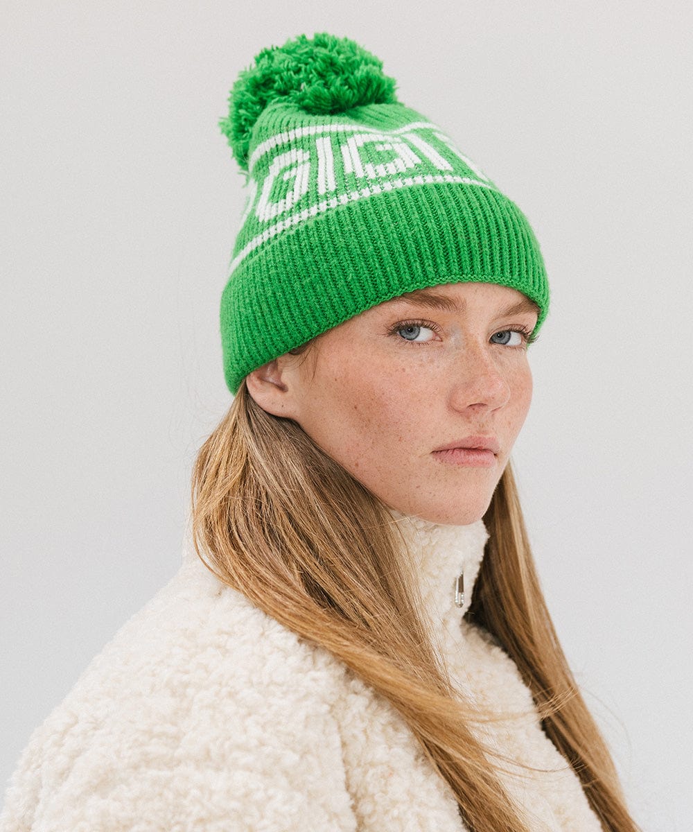 Woman wearing an evergreen colored knit beanie with pom-pom and 'GIGI PIP' branding and a white sweater against a plain background #color_evergreen
