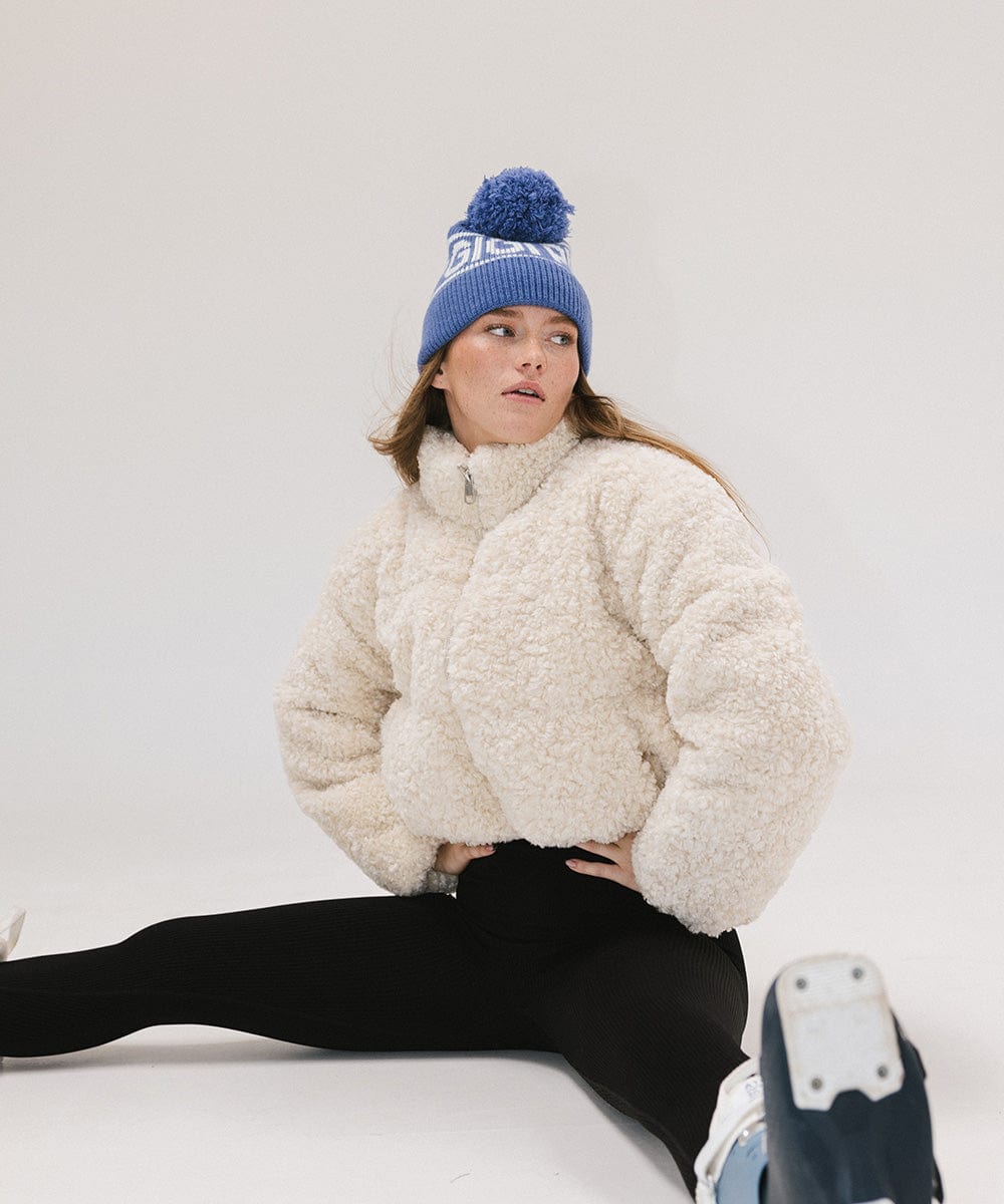 Woman wearing an arctic blue colored knit beanie with pom-pom and 'GIGI PIP' branding, a white sweater and black pants, sitting against a plain background #color_arctic blue