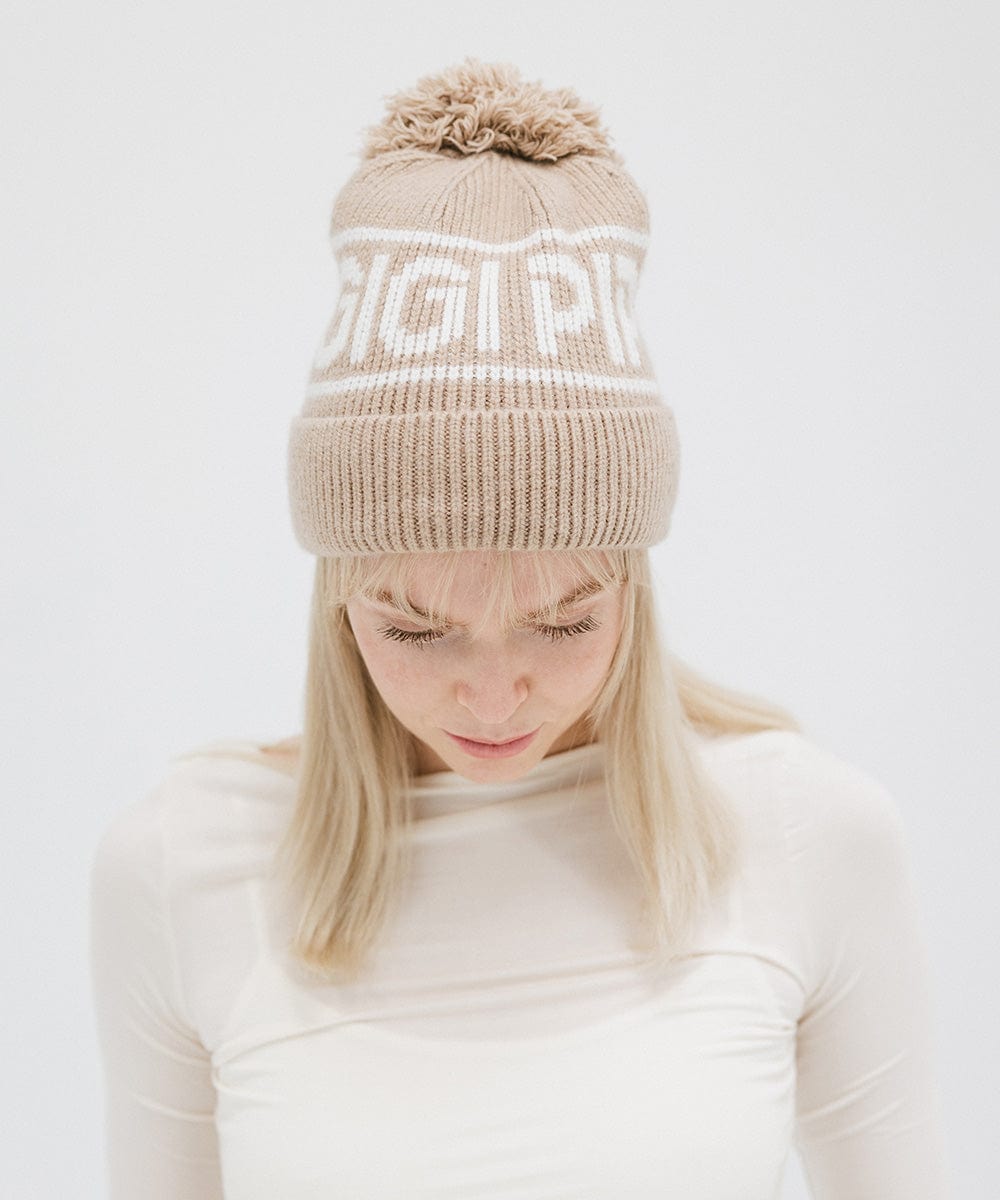 Woman wearing a taupe colored knit beanie with pom-pom and 'GIGI PIP' branding and a white top, looking down against a plain background #color_taupe