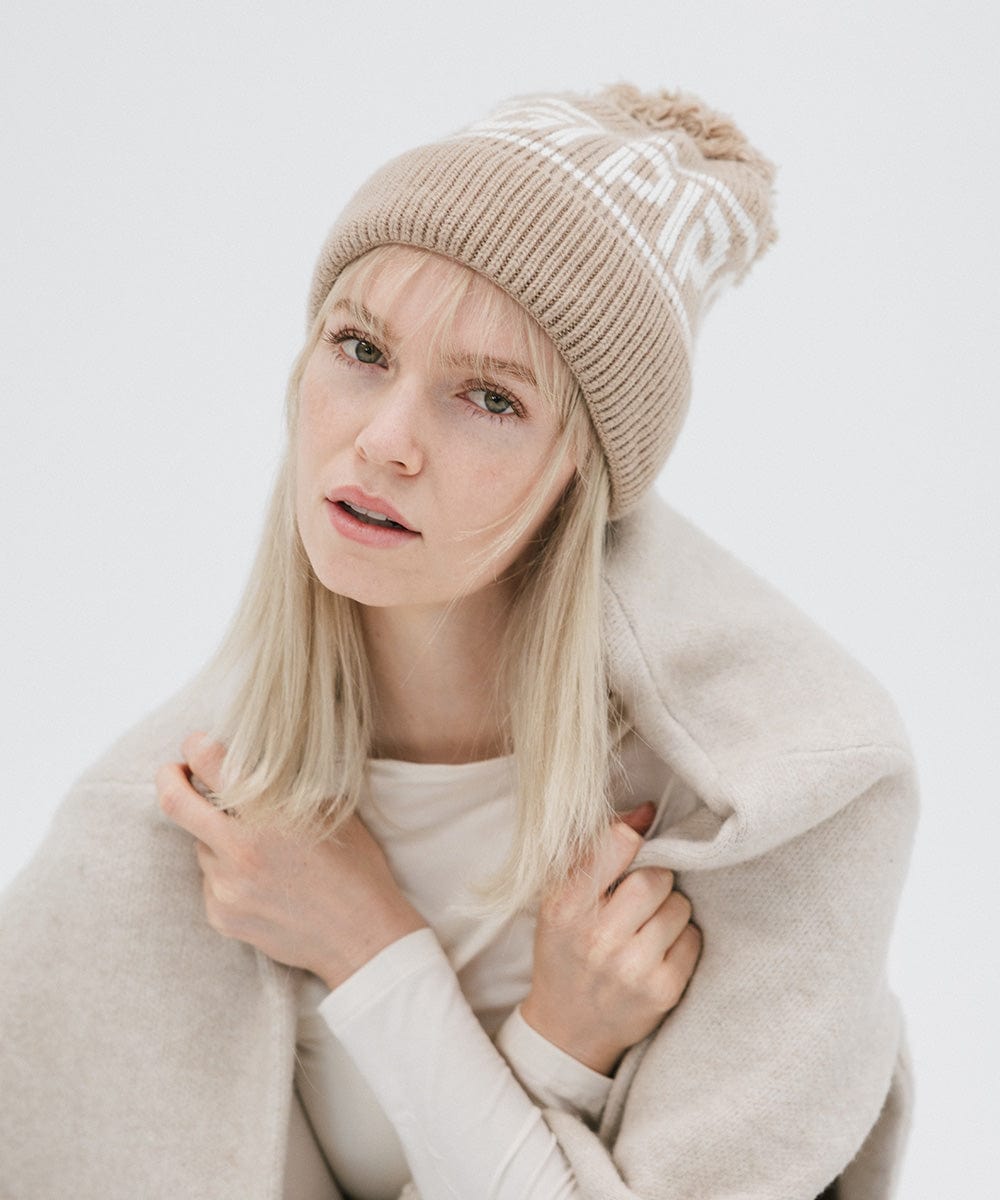 Woman wearing a taupe colored knit beanie with pom-pom and 'GIGI PIP' branding and a white top with a white coat against a plain background #color_taupe