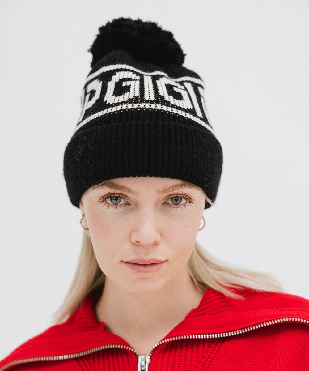 Woman wearing a black knit beanie with pom-pom and 'GIGI PIP' branding and a red jacket against a plain background #color_black