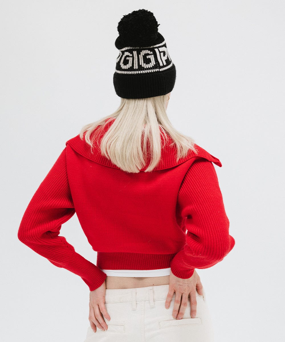 Woman wearing a black knit beanie with pom-pom and 'GIGI PIP' branding, a red jacket and white pants, facing behind against a plain background #color_black