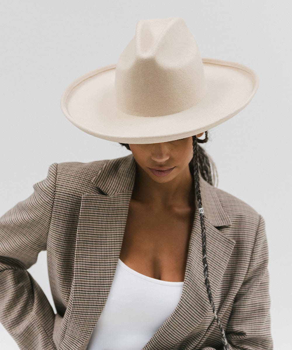 Woman wearing an off white western hat with a pencil brim and a tall crown and a white top with a coat against a plain background #color_off white