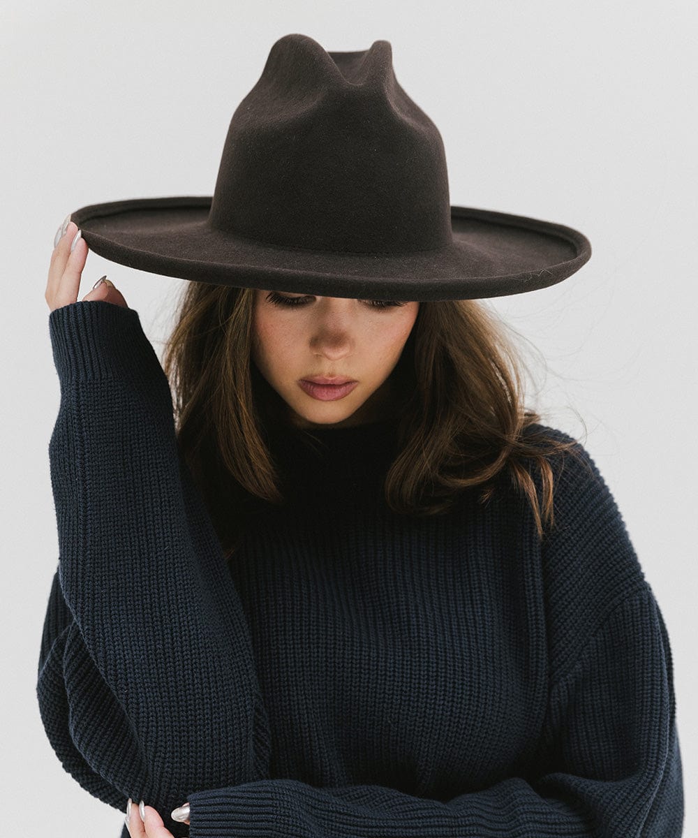 Woman wearing a dark brown western hat with a pencil brim and a tall crown and a dark blue sweater against a plain background #color_dark brown