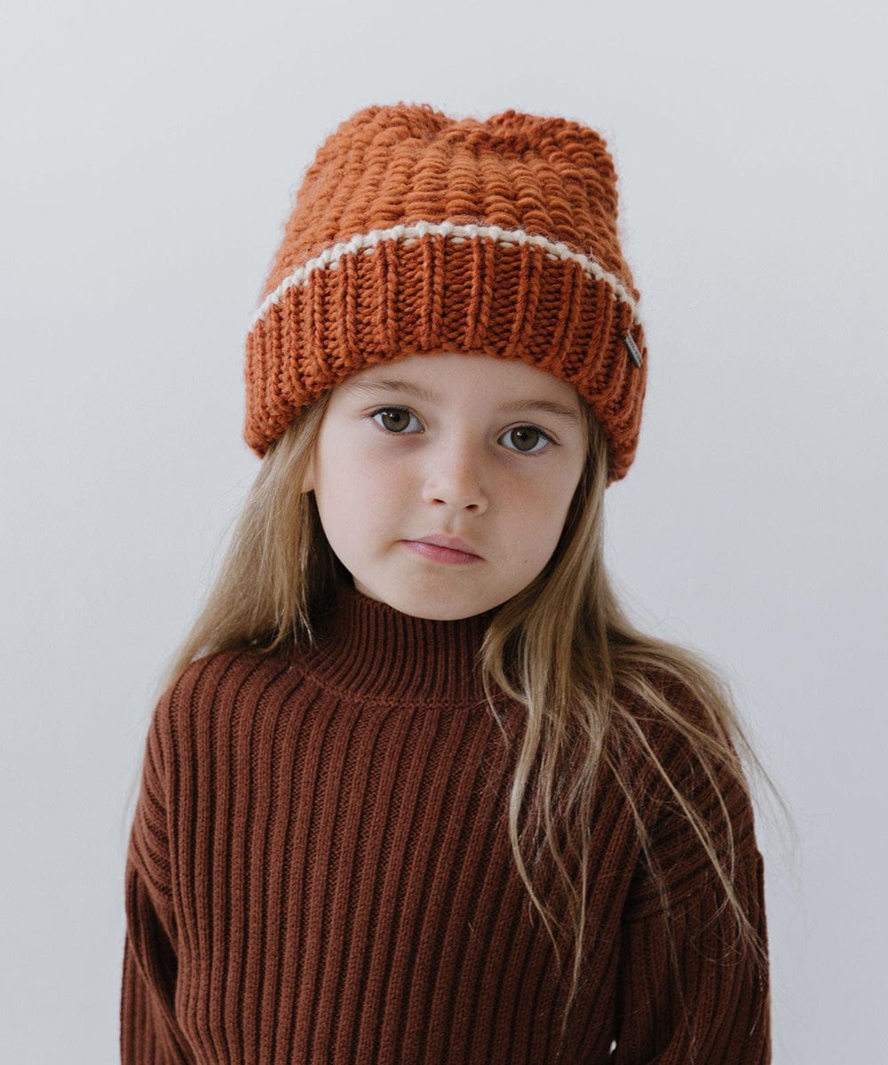 Child wearing a burnt orange vail beanie with a cream stripe and a brown sweater against a plain background #color_burnt orange-cream