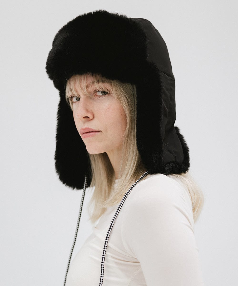 Woman wearing a black fur-trimmed hat and a white top, posing sideways against a plain background #color_black