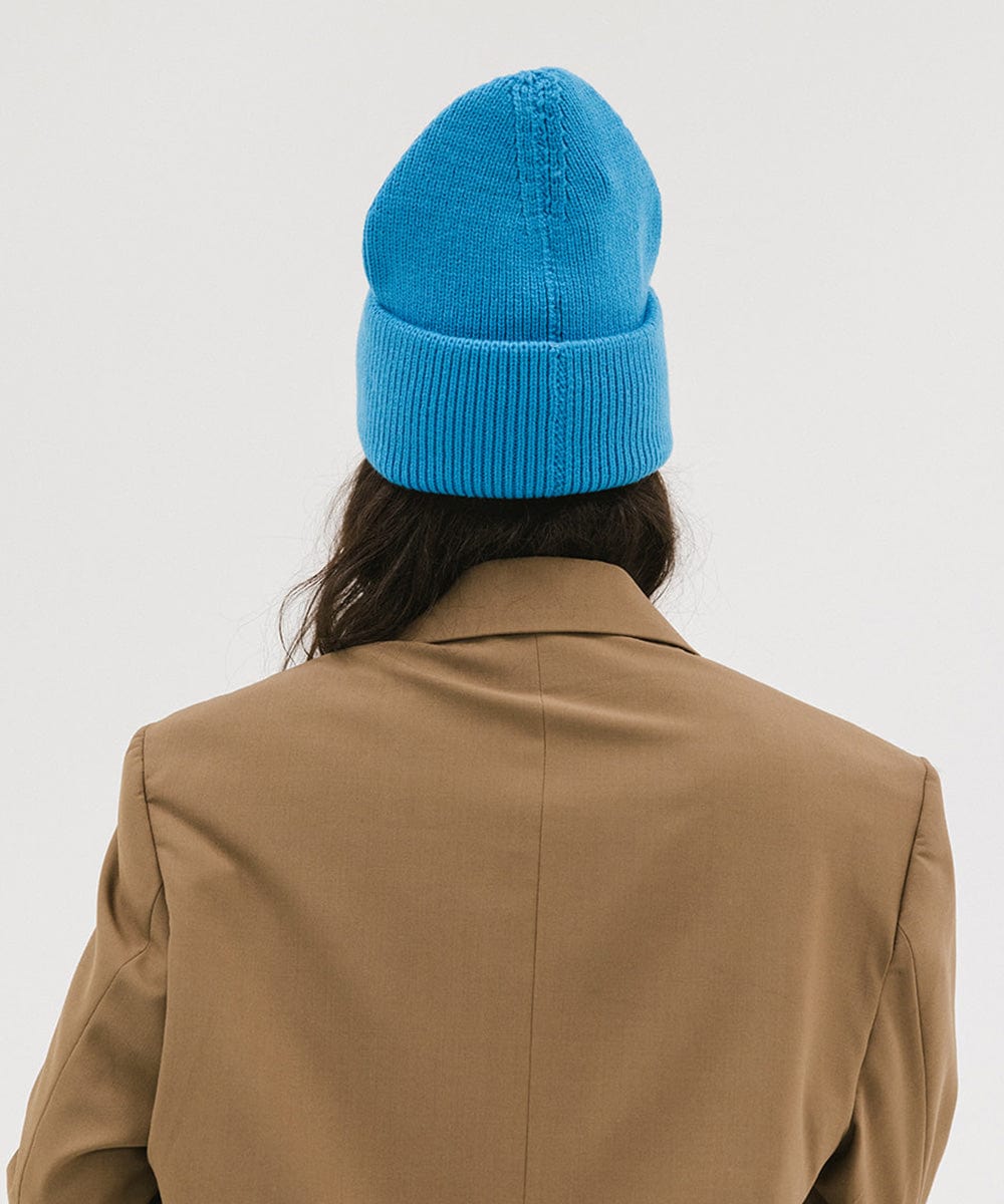 Person wearing a blue oversized knit beanie and a brown coat against a plain background #color_deja blue