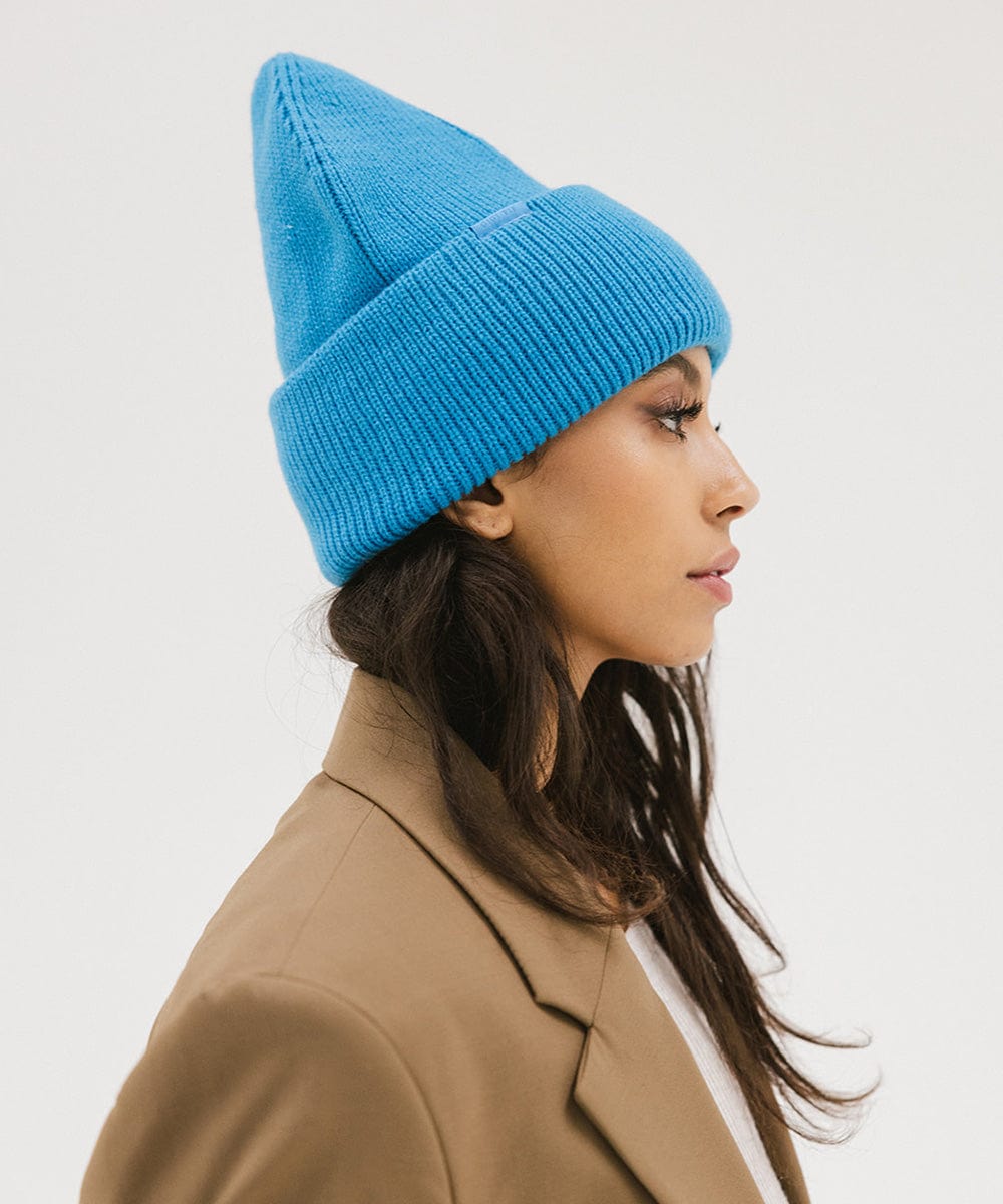 Woman wearing a blue oversized knit beanie and a brown coat, facing sideways against a plain background #color_deja blue