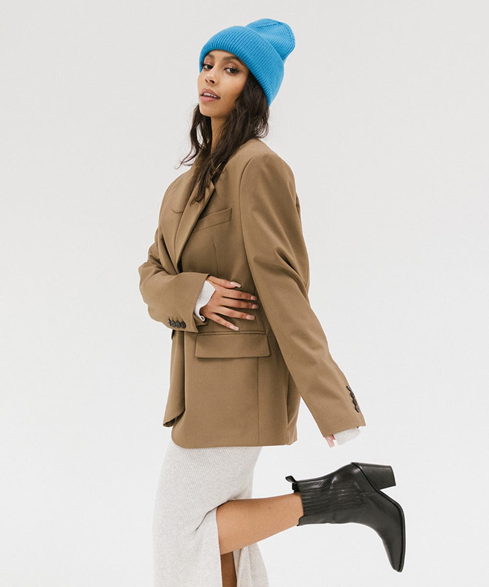 Woman wearing a blue oversized knit beanie, a brown coat, white skirt and black boots against a plain background #color_deja blue